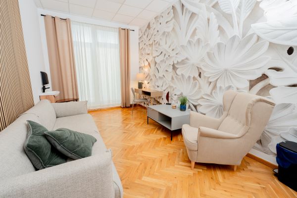 Gabinet psychologiczny. Szara kanapa z lewej strony, z prawej kremowy fotel. Na ścianie tapeta we wzór białego kwiata. Na końcu gabinetu okno z zasłoną.