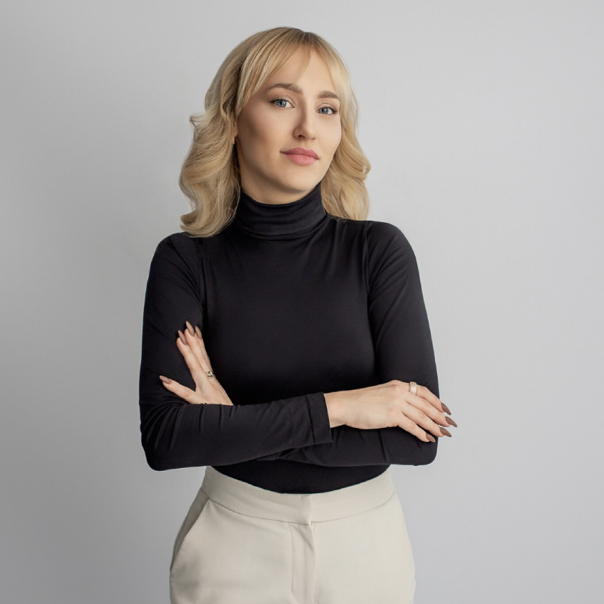 Uśmiechnięta kobieta w blond włosach, czarnym swetrze oraz w kremowych spodniach.