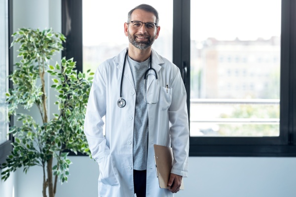 Uśmiechnięty lekarz w okularach oraz w białym kitlu, na tle oszklonej ściany. Z boku zielony kwiat