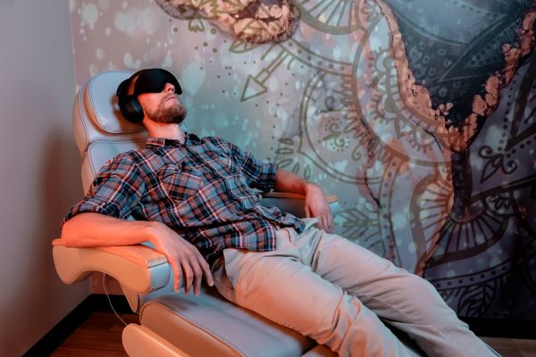 Mężczyzna, z założoną opaską na oczy oraz słuchawkami leży na szarym fotelu. W tle kolorowa ściana.